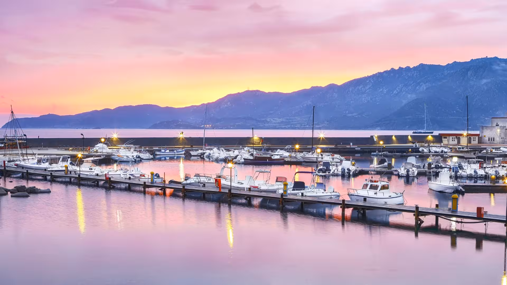 Serene marina in Villasimius at sunset, with boats docked and mountains in the background.