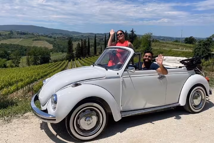 Smiling duo waving from a white vintage Beetle amidst the rolling vineyards of Tuscany.