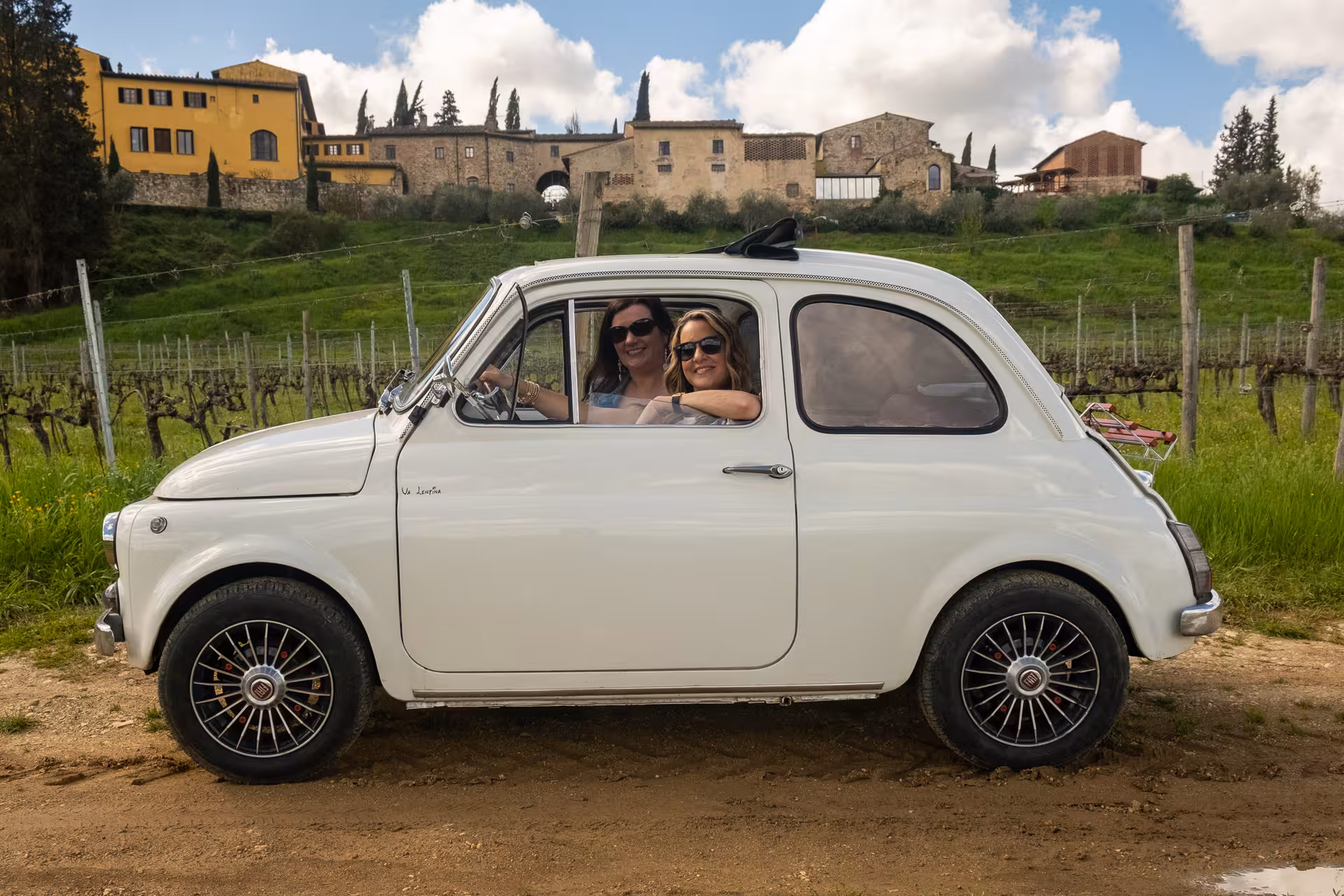 Two women smiling inside a vintage FIAT 500 parked near vineyards on a Chianti Classico wine tour.
