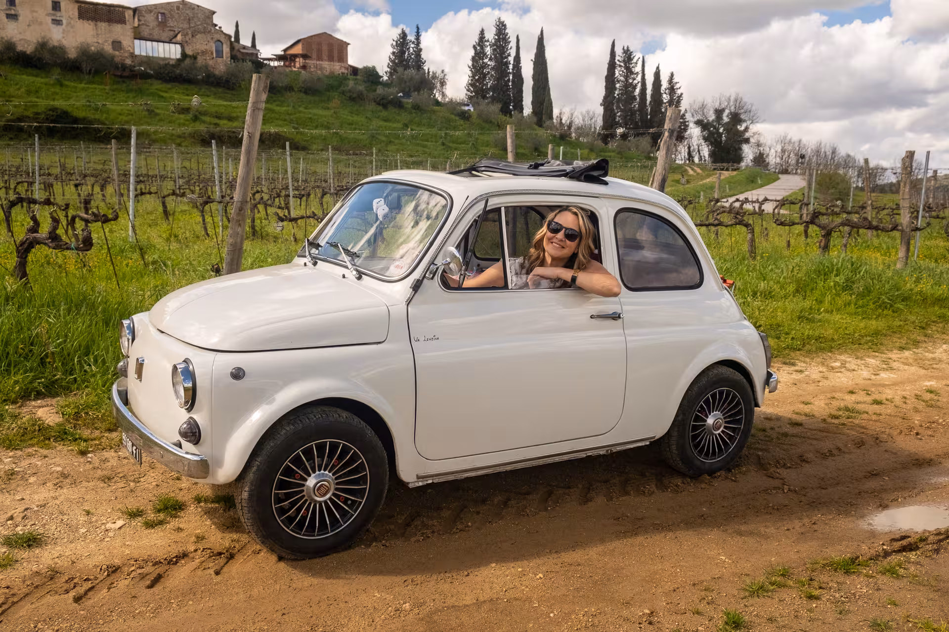 Smiling woman in a vintage white FIAT 500 driving through scenic vineyards in Tuscany on a sunny day.