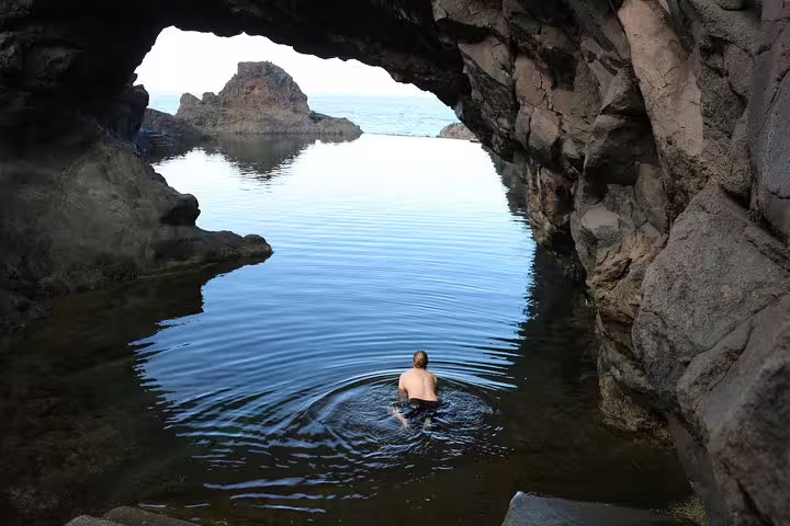 Adventurer enjoys a refreshing swim in a tranquil volcanic pool beneath a rugged rock archway, perfect for exploration.