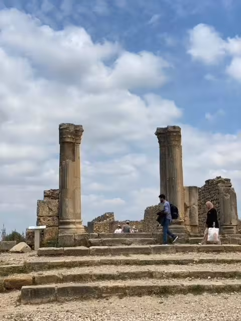 Visitors explore the ancient Roman ruins of Volubilis, featuring iconic stone columns under a partly cloudy sky.