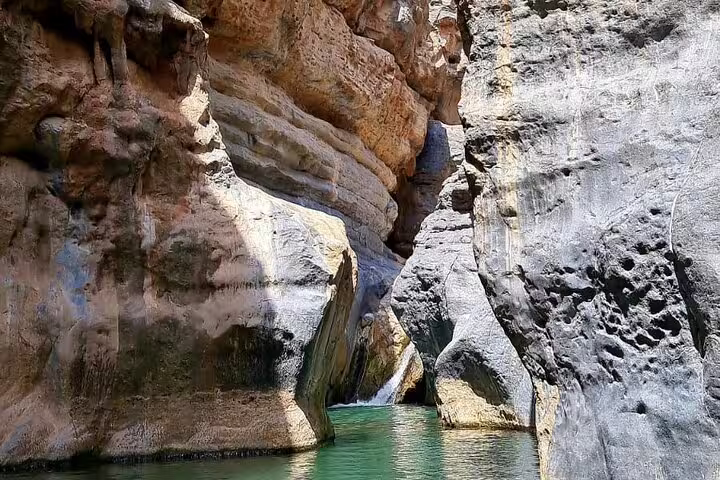 Narrow, sunlit canyon passage in Wadi Dham with emerald waters and towering rock walls showcasing Oman's natural wonders.
