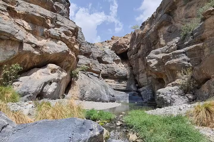 Serene Wadi Dham landscape with rugged rock formations, lush greenery, and tranquil water reflecting sunny skies in Oman.