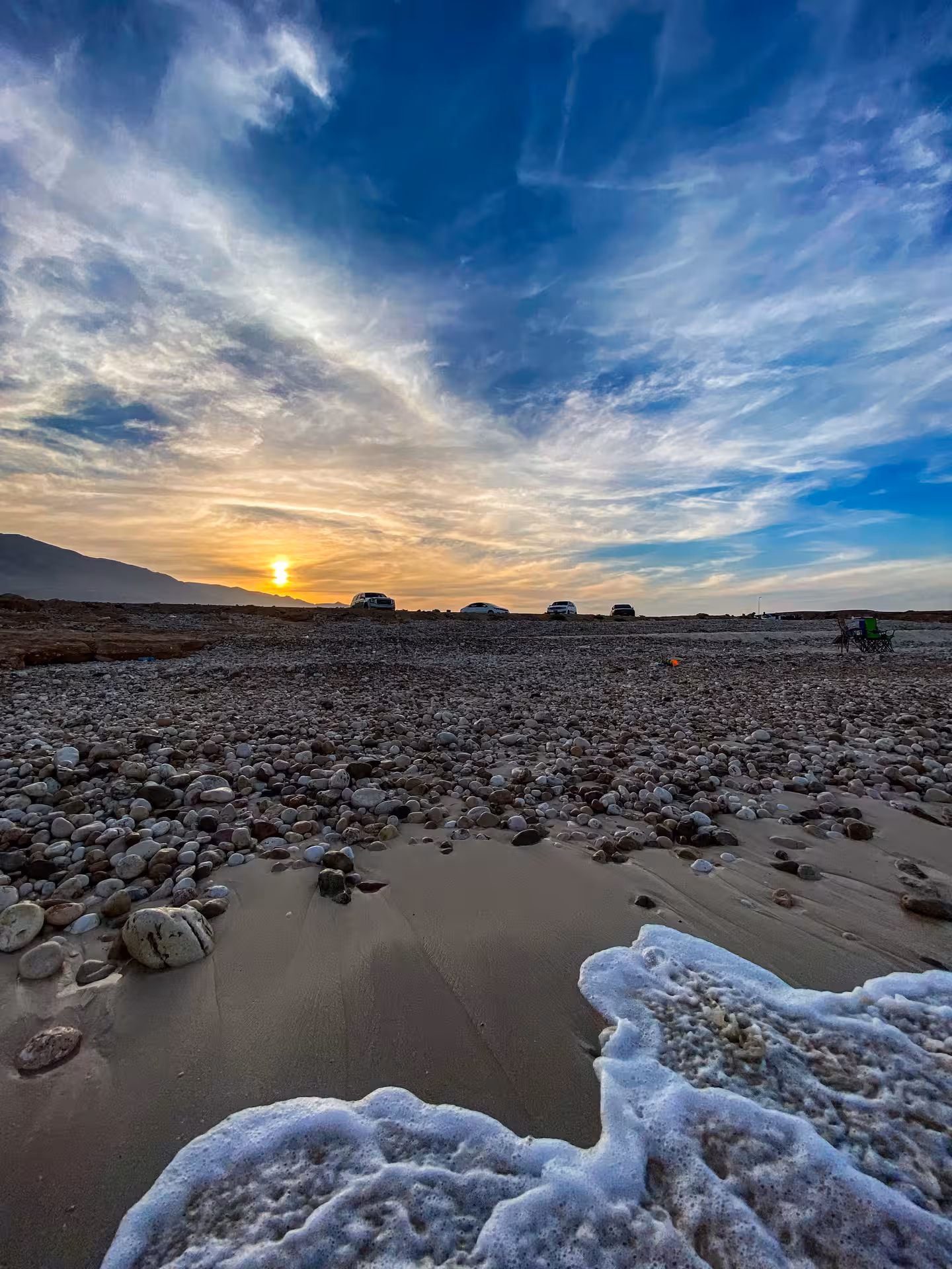 Foamy waves wash over a pebbled Oman beach at sunset, with tour vehicles parked along the shore near Wadi Shab