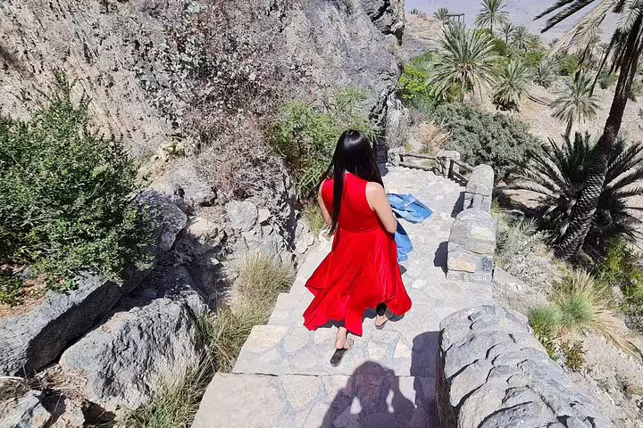 Woman in red dress walking down stone steps amidst rocky terrain and palm trees in Wakan Village.