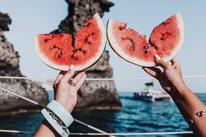 Refreshing watermelon slices held against the backdrop of Santorini's rocky coastline on a catamaran cruise.