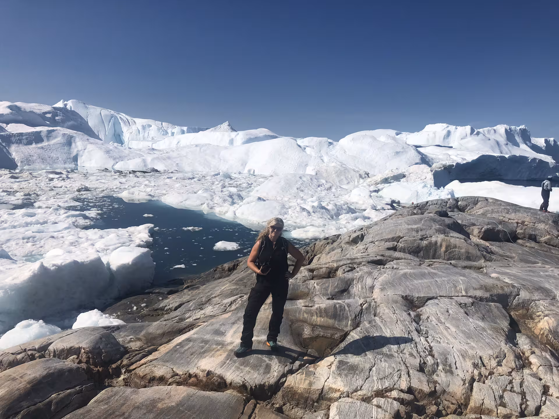 Traveler exploring stunning ice-covered landscapes and rocky terrain in West Greenland under a clear blue sky.