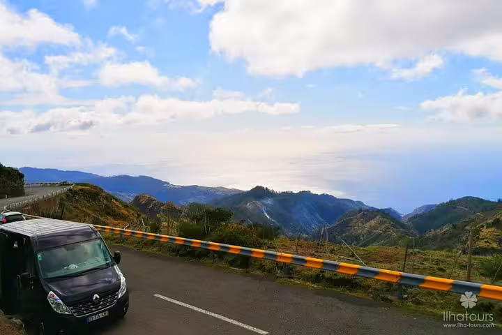 Scenic mountain view with a tour van on a winding road overlooking the ocean under a blue sky.