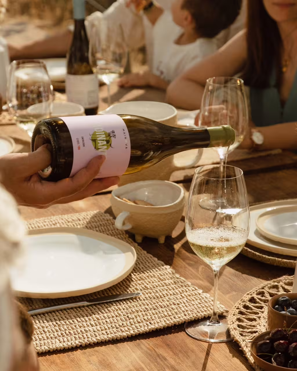 A hand pours white wine into a glass at an outdoor tasting event, surrounded by plates and bowls on a rustic table setting.