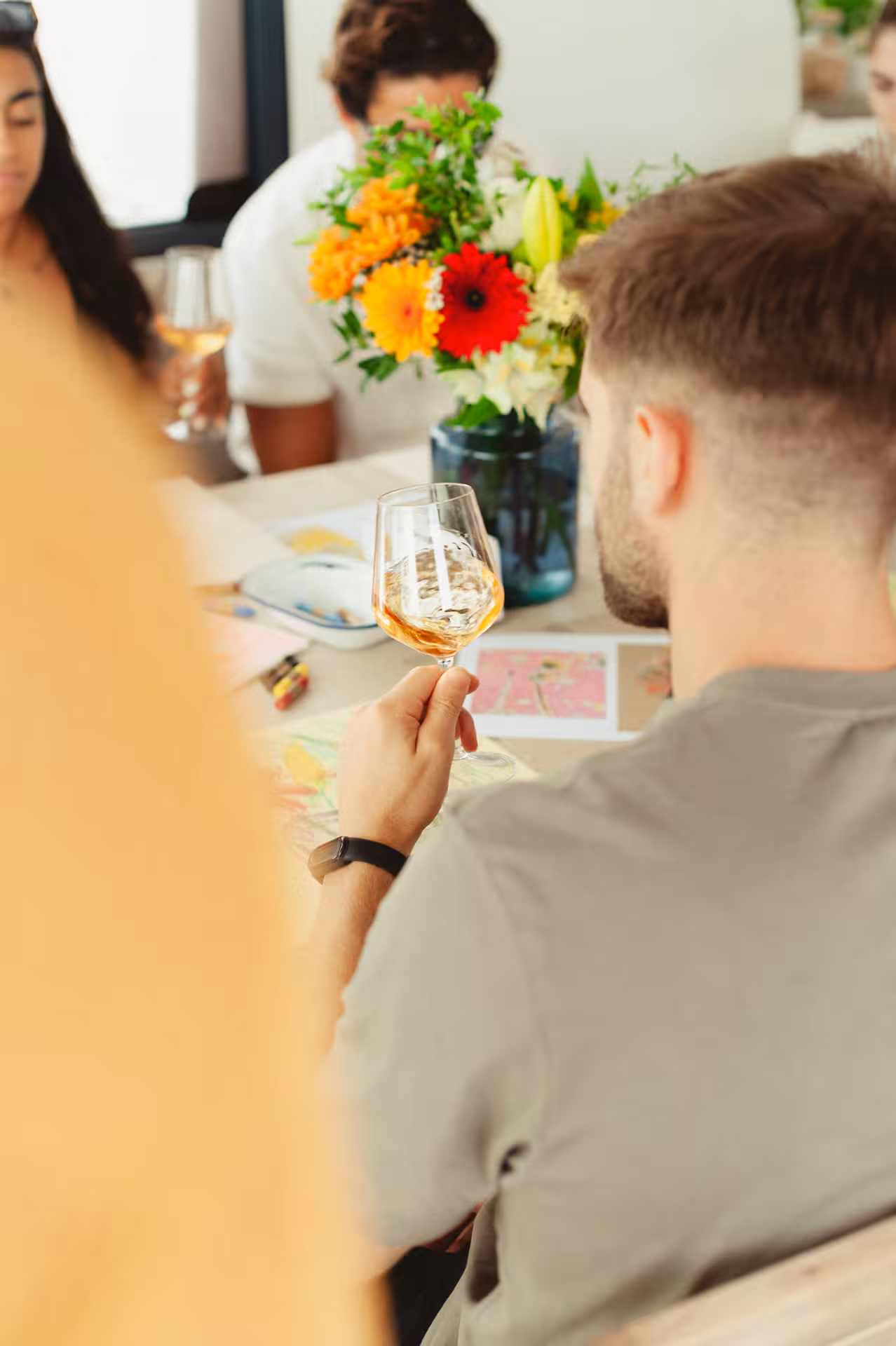 Participant sipping white wine while painting at a Wine & Art class, with bright floral centerpiece on table
