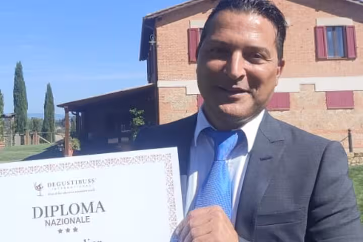 Man in suit holding a diploma certificate outside a rustic building in Rome, promoting wine education course.