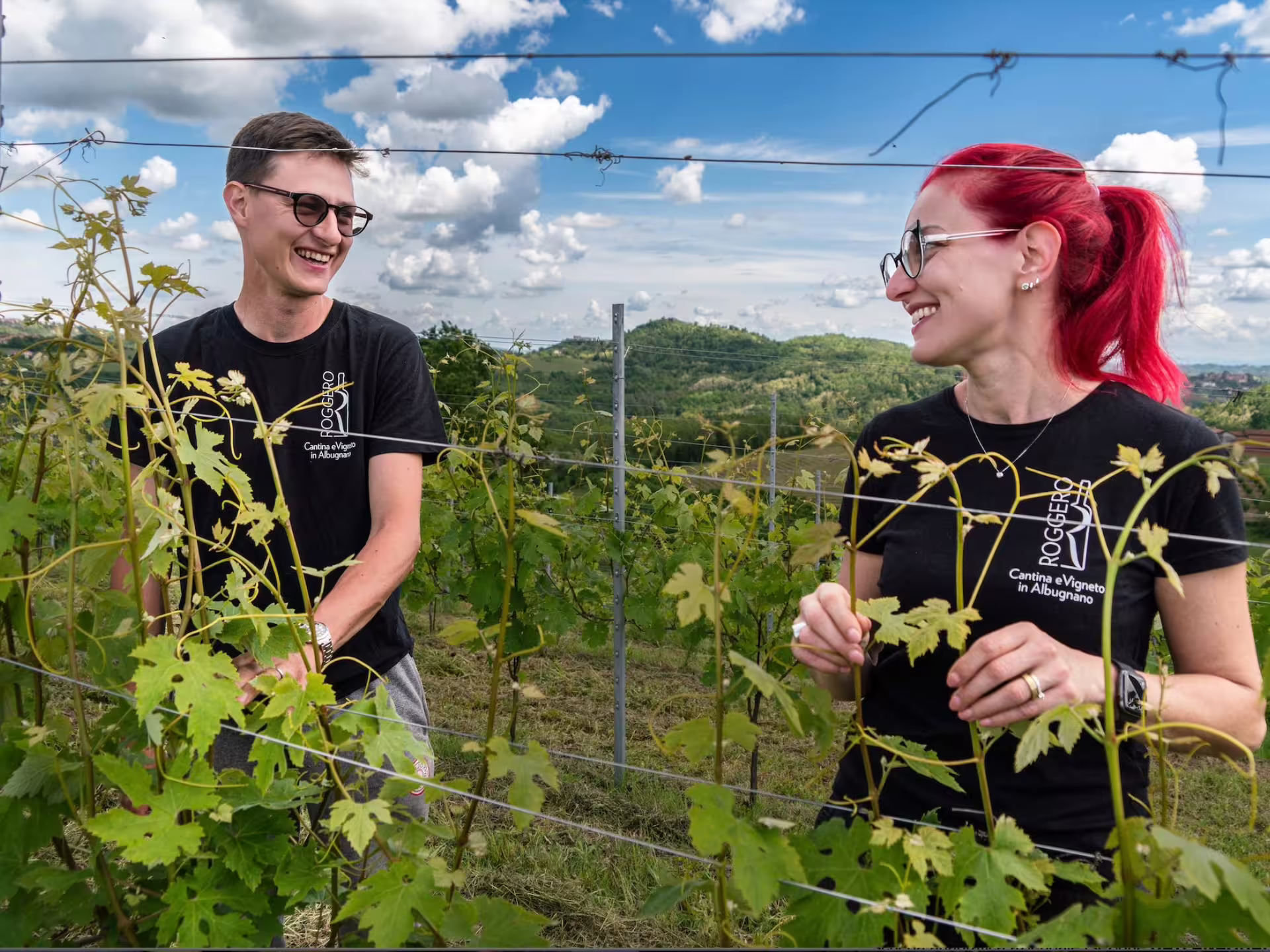 Two people smiling and tending to grapevines in a picturesque vineyard near Asti, Italy.