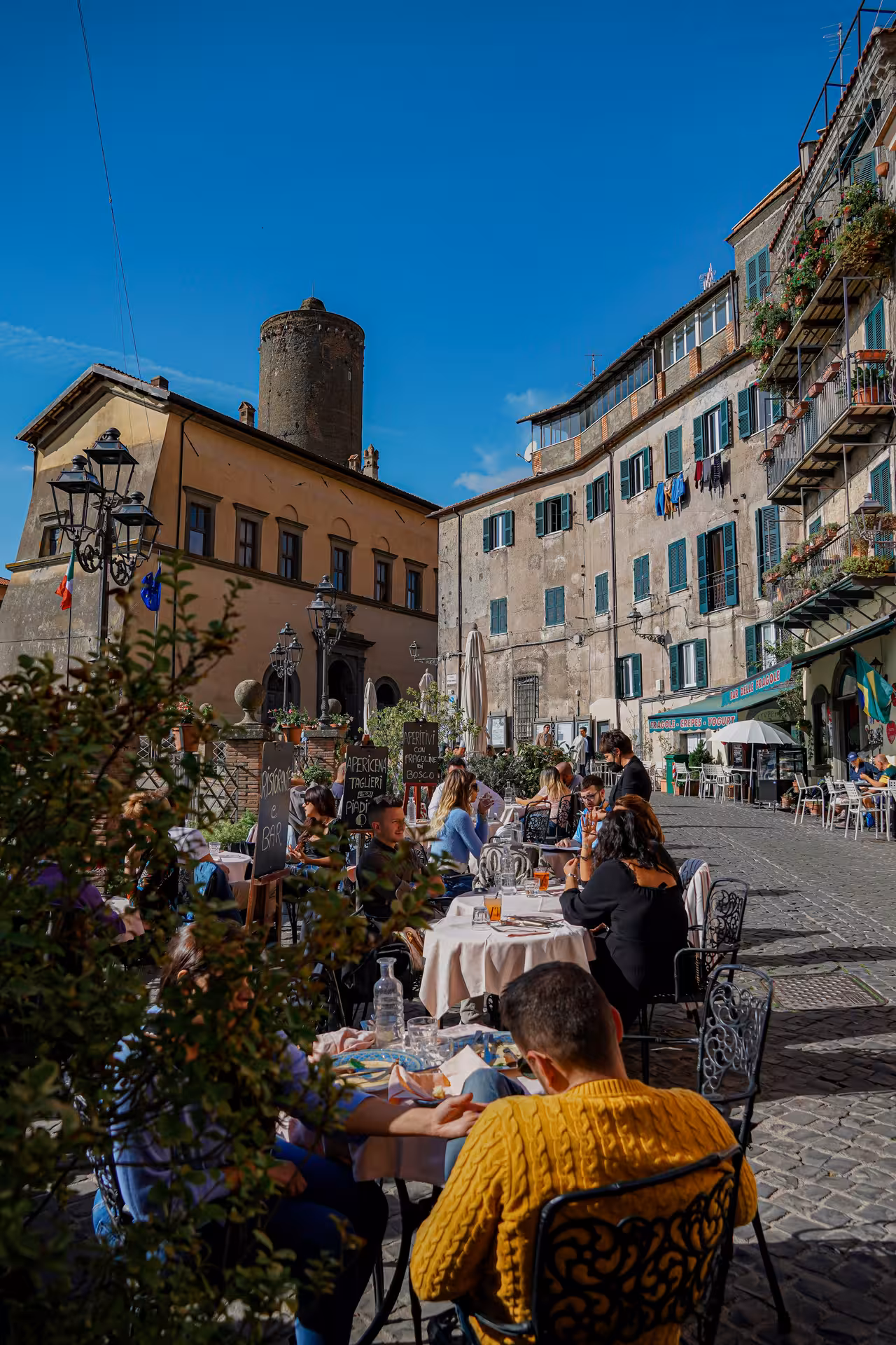 Outdoor dining in a picturesque Italian square south of Rome, bustling with people enjoying meals under clear skies.