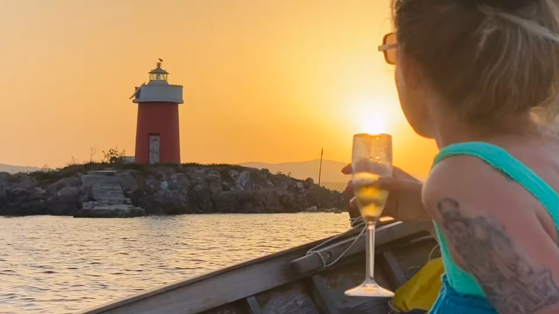 Woman enjoying aperitif on a sunset sailing tour near Alghero lighthouse, creating a serene coastal experience.