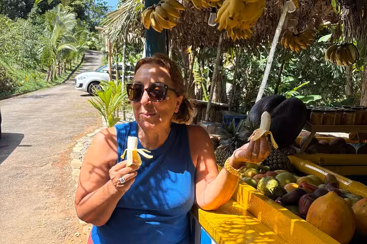 Woman savoring a fresh banana at a roadside fruit stand on a sunny Mahe Island tour.