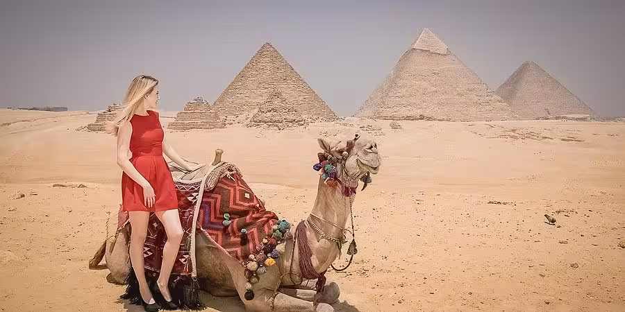 Woman in red dress standing beside a decorated camel with the Pyramids of Giza in the background under a clear sky.