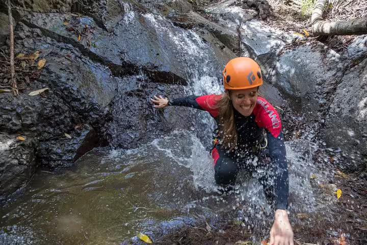 Adventurous woman enjoys canyoning under a small waterfall in the lush rainforest, wearing a helmet and wetsuit.