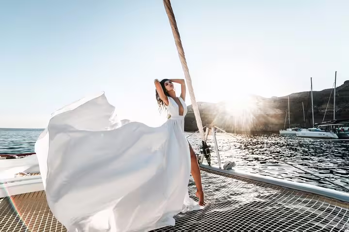 Woman enjoying the breeze on a catamaran in Santorini with a flowing white dress and scenic ocean view.