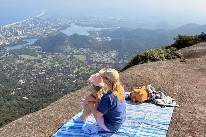 Woman with child enjoying a scenic picnic on a rock overlooking stunning Tijuca Forest and city views below.