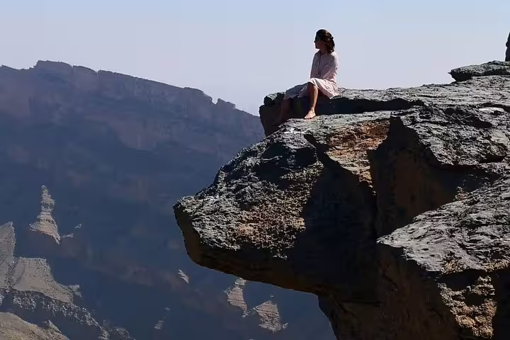 Woman sitting on the edge of a dramatic cliff at Jebel Shams, overlooking a vast canyon landscape during sunset.