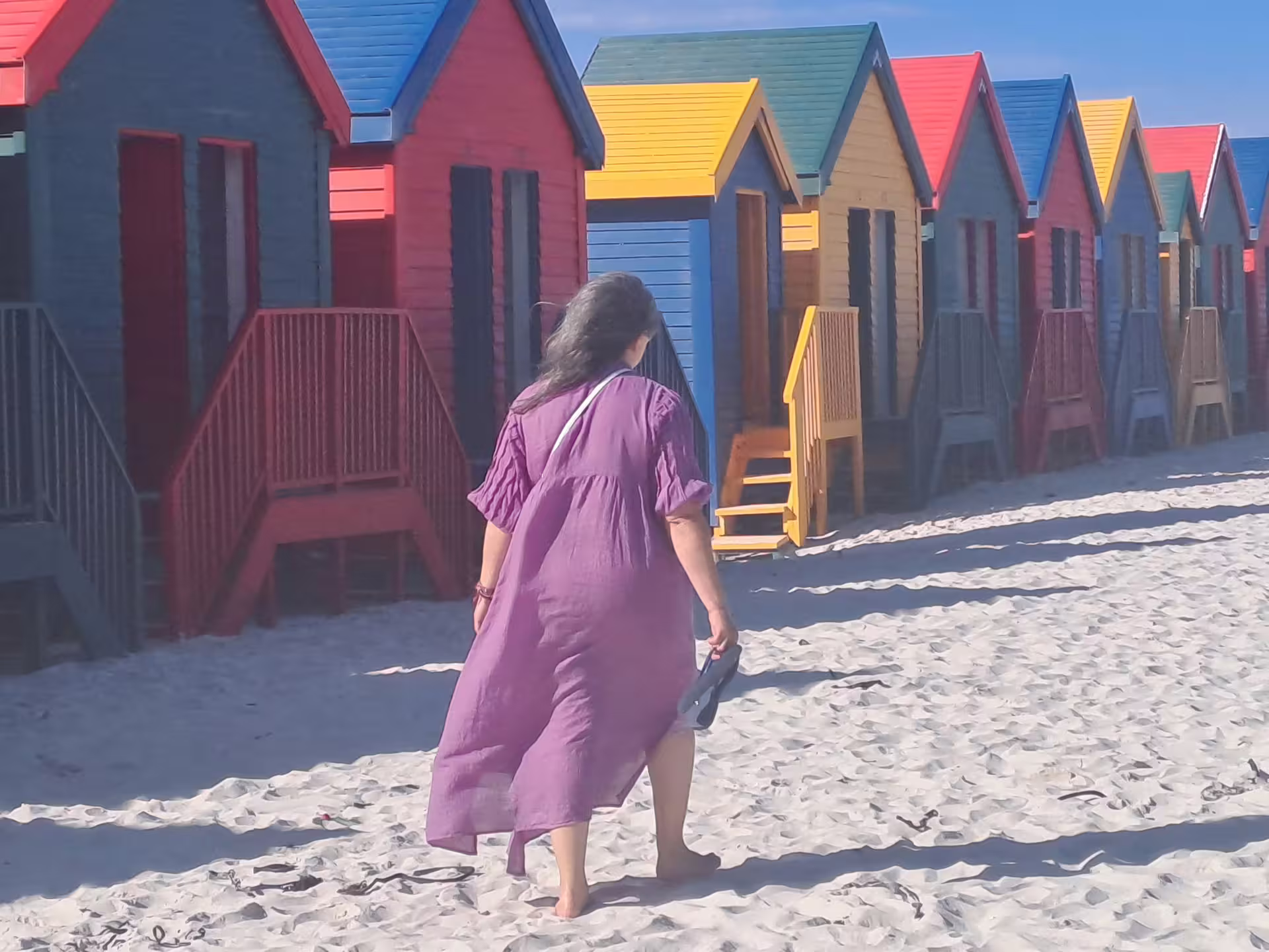 Woman walks along the sandy beach in front of colorful beach huts in Cape Town.