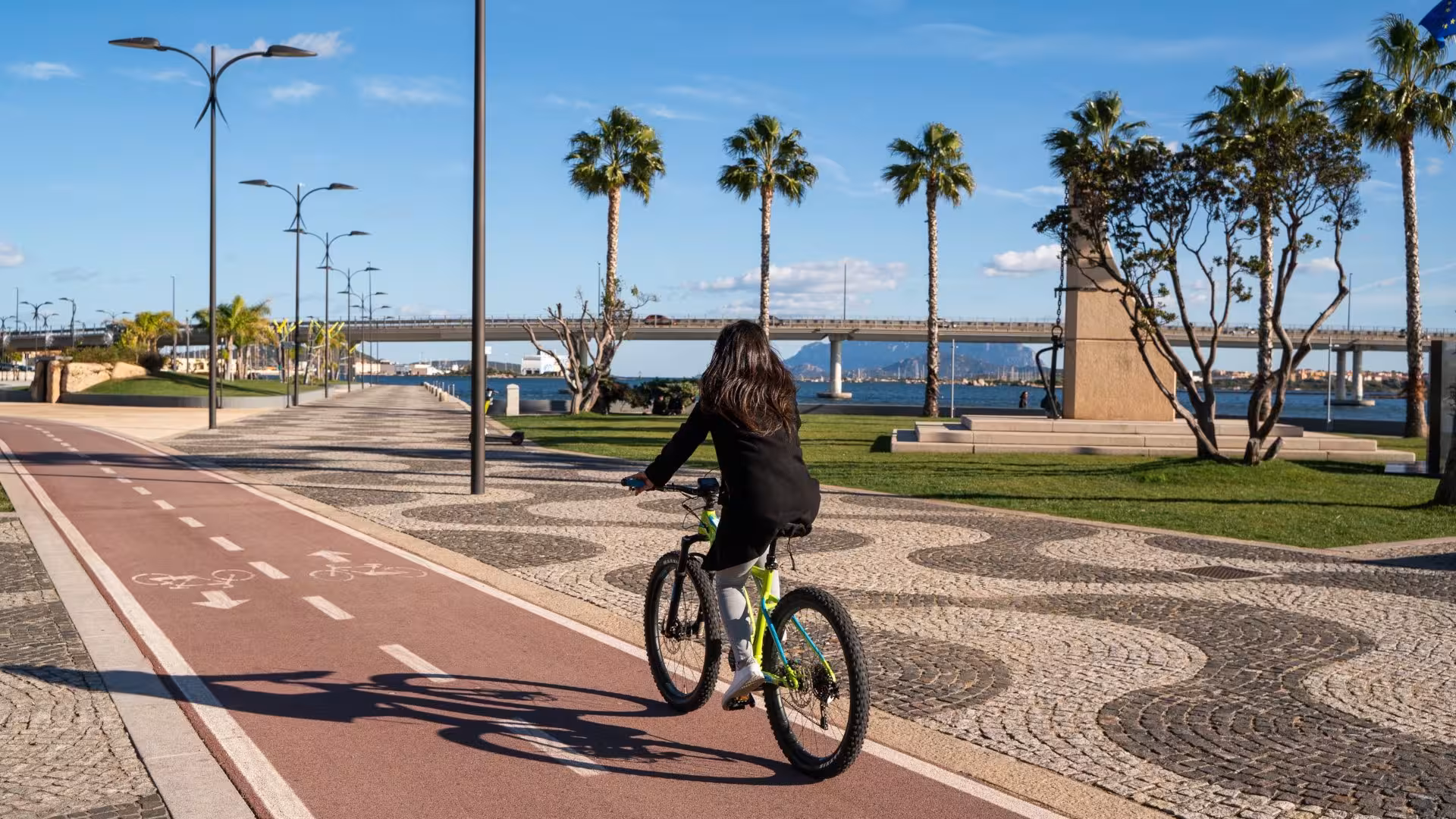 Woman enjoying a scenic ride on an e-bike along Olbia's coastal path in Sardinia, surrounded by palm trees and sea views.