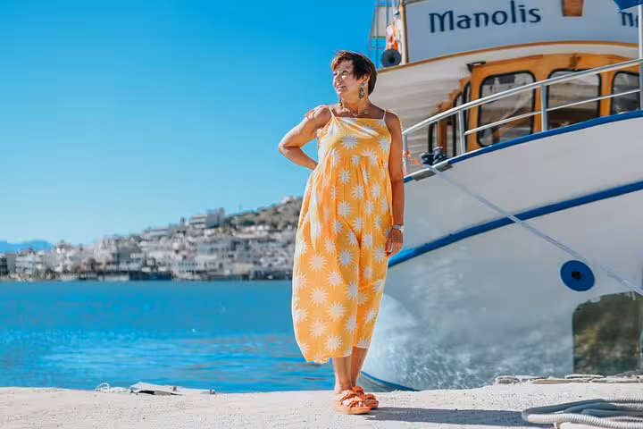 Woman in floral dress poses by the sea with a boat in Elounda, perfect for a private photoshoot experience.