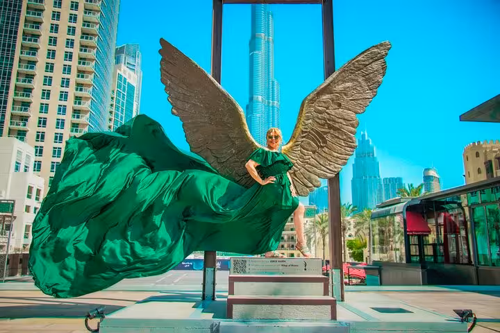 Woman in a flowing green dress poses with angel wings, Burj Khalifa towering in the background, perfect for Instagram.