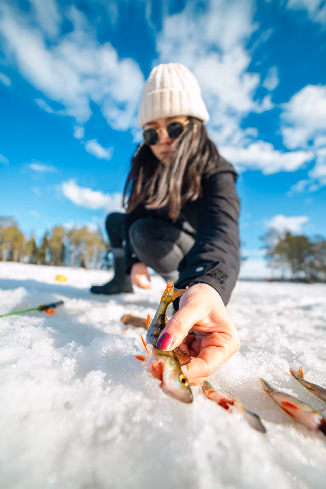 Woman in warm attire holds small fish on snowy ice, showcasing a hands-on ice fishing experience in bright sunlight.