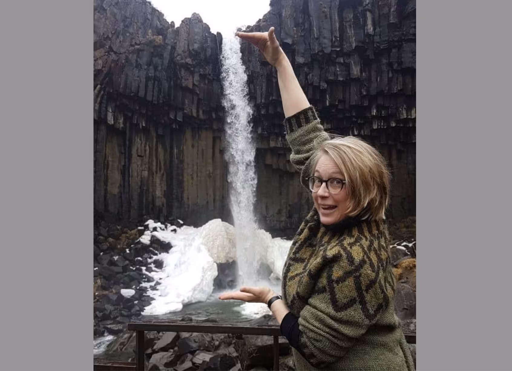 Smiling woman in Icelandic sweater playfully poses with Svartifoss waterfall during winter knitting tour.