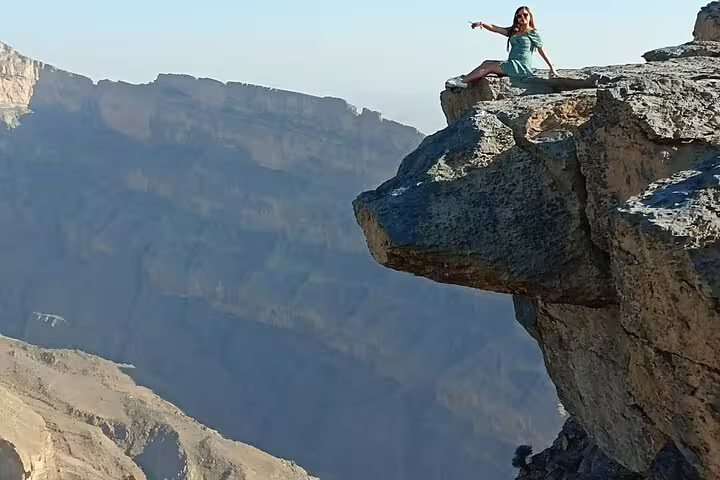 Adventurous woman sits on dramatic rocky ledge overlooking breathtaking Jebel Shams canyon during overnight camping tour.