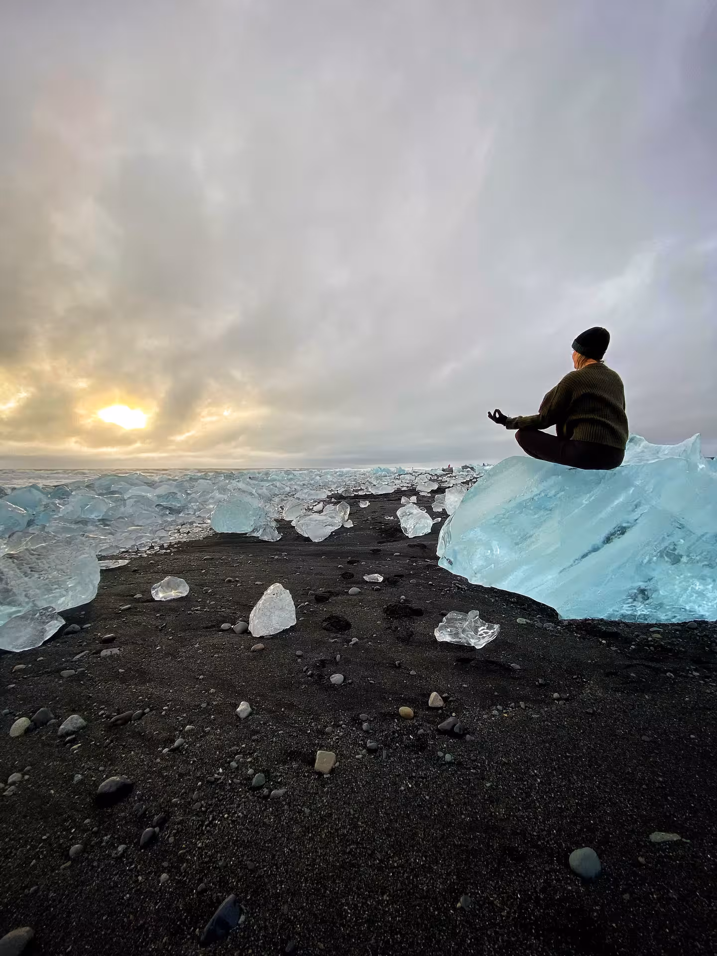 Woman meditating on an ice block at sunset on a black sand beach during a winter hiking tour in Iceland.