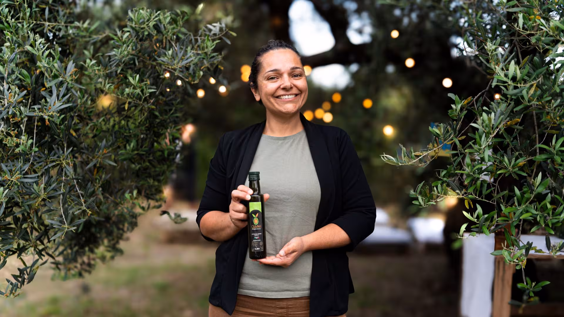 Smiling woman holding a bottle of olive oil amidst illuminated olive trees in Lotzorai, Tortolì oil mill tour.