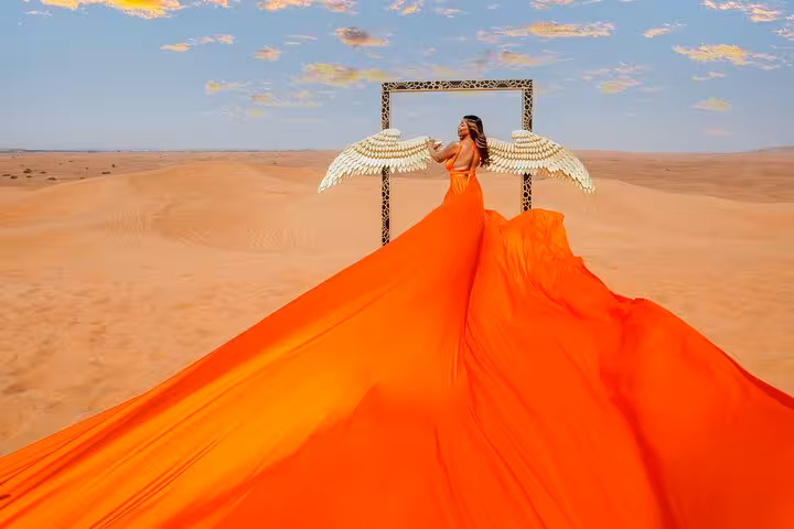 Woman in vibrant orange dress with angel wings stands in Dubai desert for Flying Dress Photography experience.
