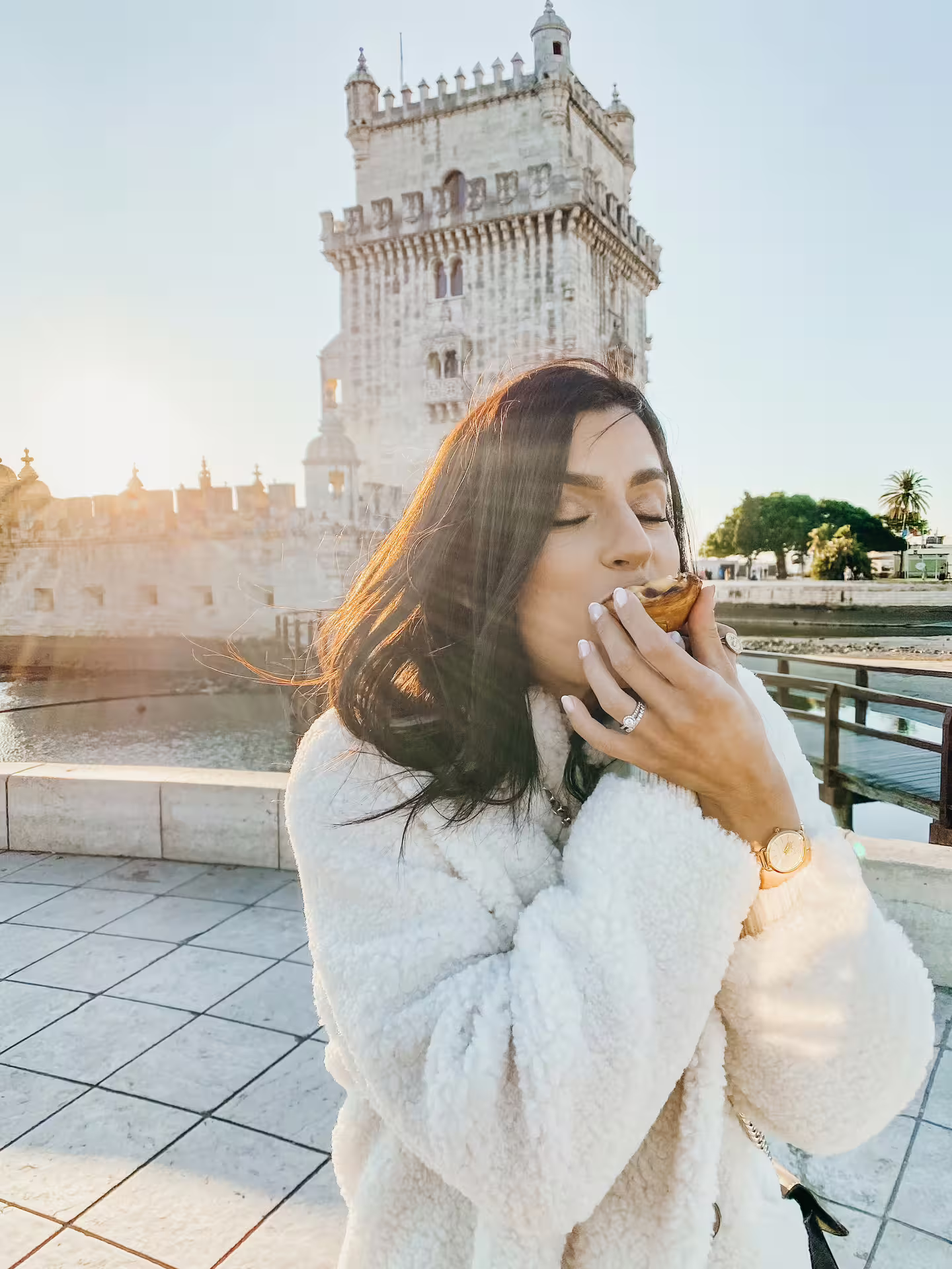 Woman enjoying a pastel de nata near Belém Tower at sunset in Lisbon.