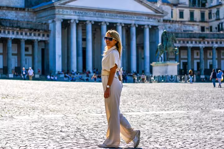 Stylish woman walks confidently in front of Naples' historic Piazza del Plebiscito under sunny skies.