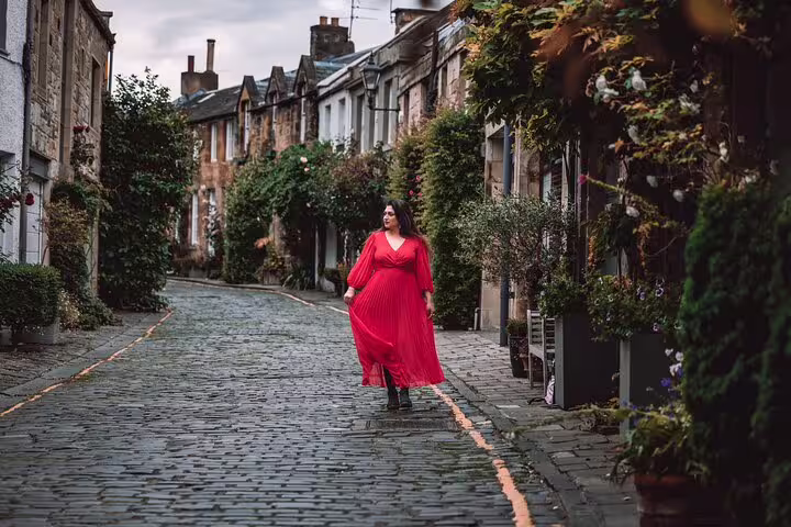 Woman in a flowing red dress strolls along a charming cobblestone street in Edinburgh lined with lush greenery.