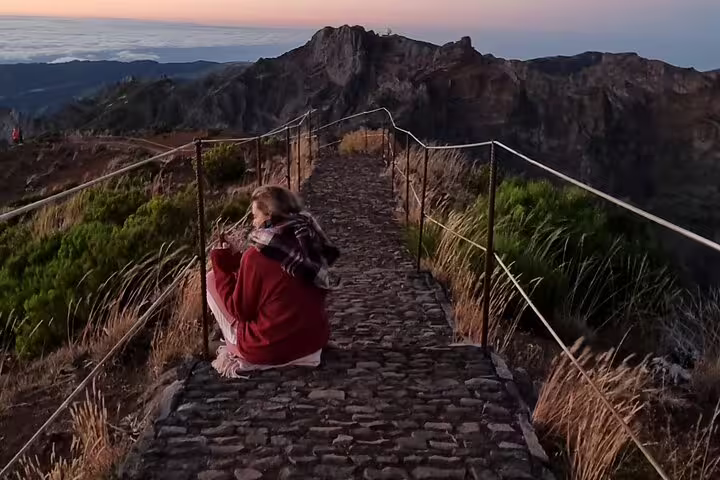 Woman in red sweater enjoys breathtaking sunrise view from Areeiro Peak, Madeira, surrounded by rugged mountain landscape.