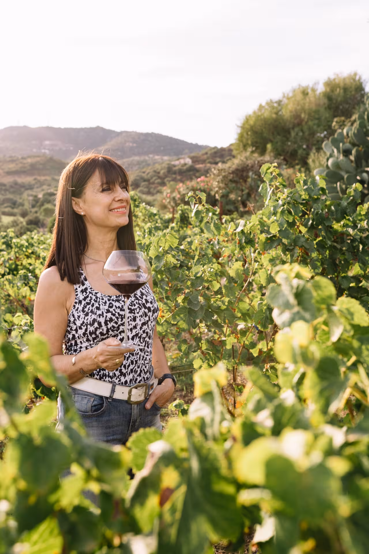 Woman enjoying a glass of red wine amidst lush green vineyards with scenic mountain views in Gallura, Olbia.