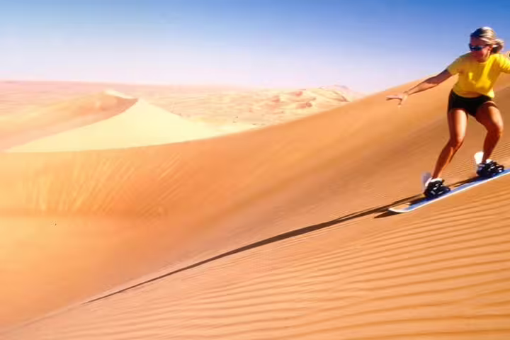 A woman sandboarding down a vast desert dune under a clear blue sky, showcasing the thrill of a Red Dunes desert safari adventure.