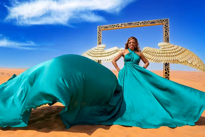 Woman in flowing teal dress with golden wings in Dubai desert during flying dress photography tour.