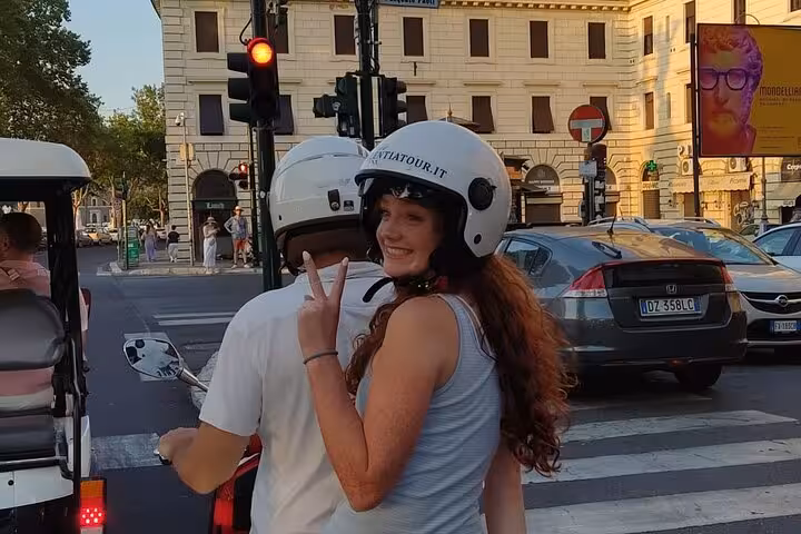 A woman in a helmet flashes a peace sign while riding a Vespa during a Rome Highlights guided tour.