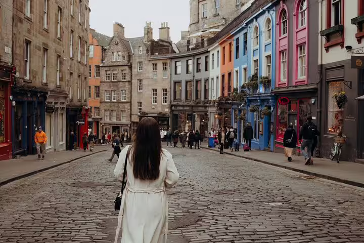 Woman in a white coat walks down Victoria Street, Edinburgh, with colorful buildings in view near Edinburgh Castle.