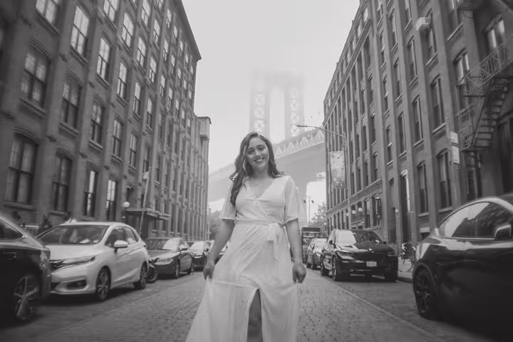 Woman in white dress stands confidently on a cobblestone street with iconic Manhattan Bridge in foggy background.