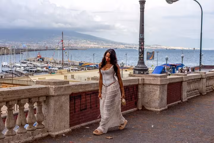Woman in a white dress walking along Naples waterfront with picturesque harbor and Mount Vesuvius in the background.