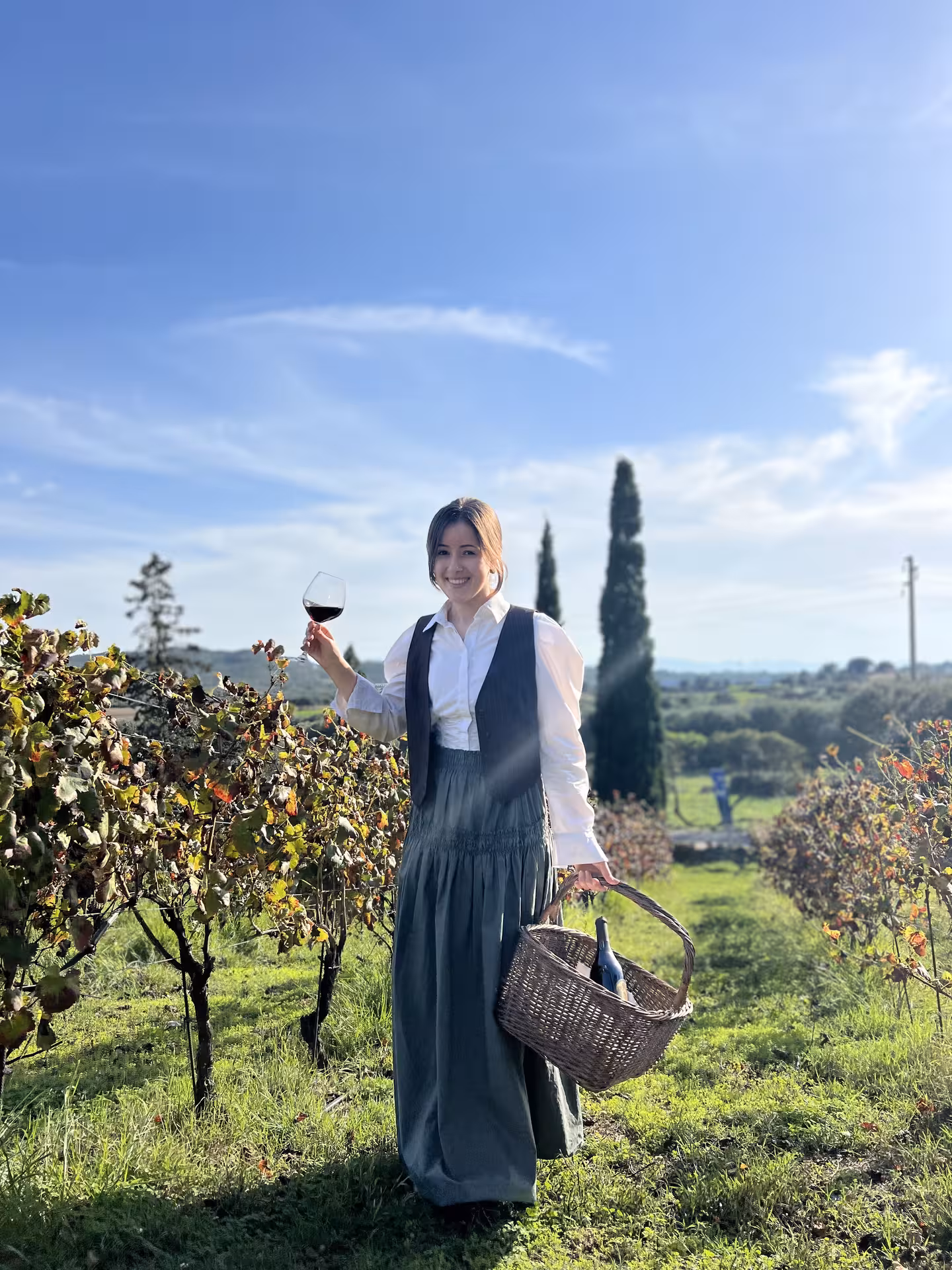 Smiling woman holding wine glass and basket, standing in lush Olbia vineyard.