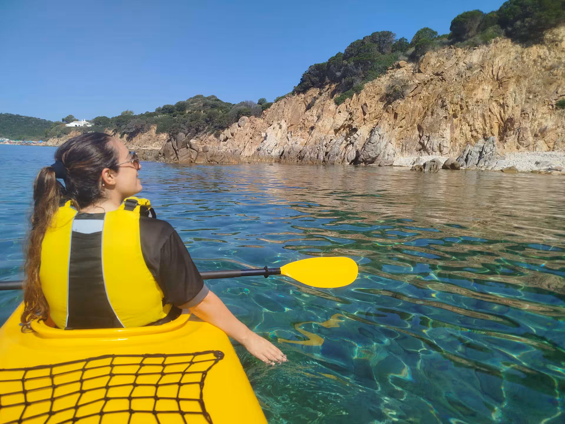 Woman in a yellow kayak enjoys the serene waters and rocky coastline of Tuerredda on a sunny day in Sardinia.