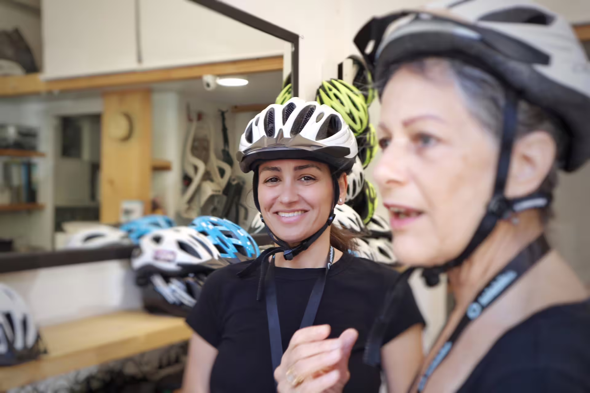 Two women wearing helmets smiling inside a bike rental shop in Athens.