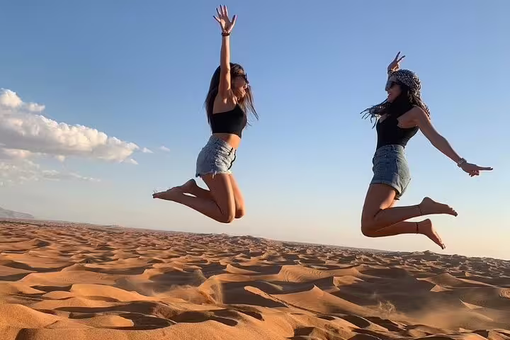 Two women joyfully leap over the vast sand dunes of Dubai's desert, capturing the thrill of a morning safari adventure.